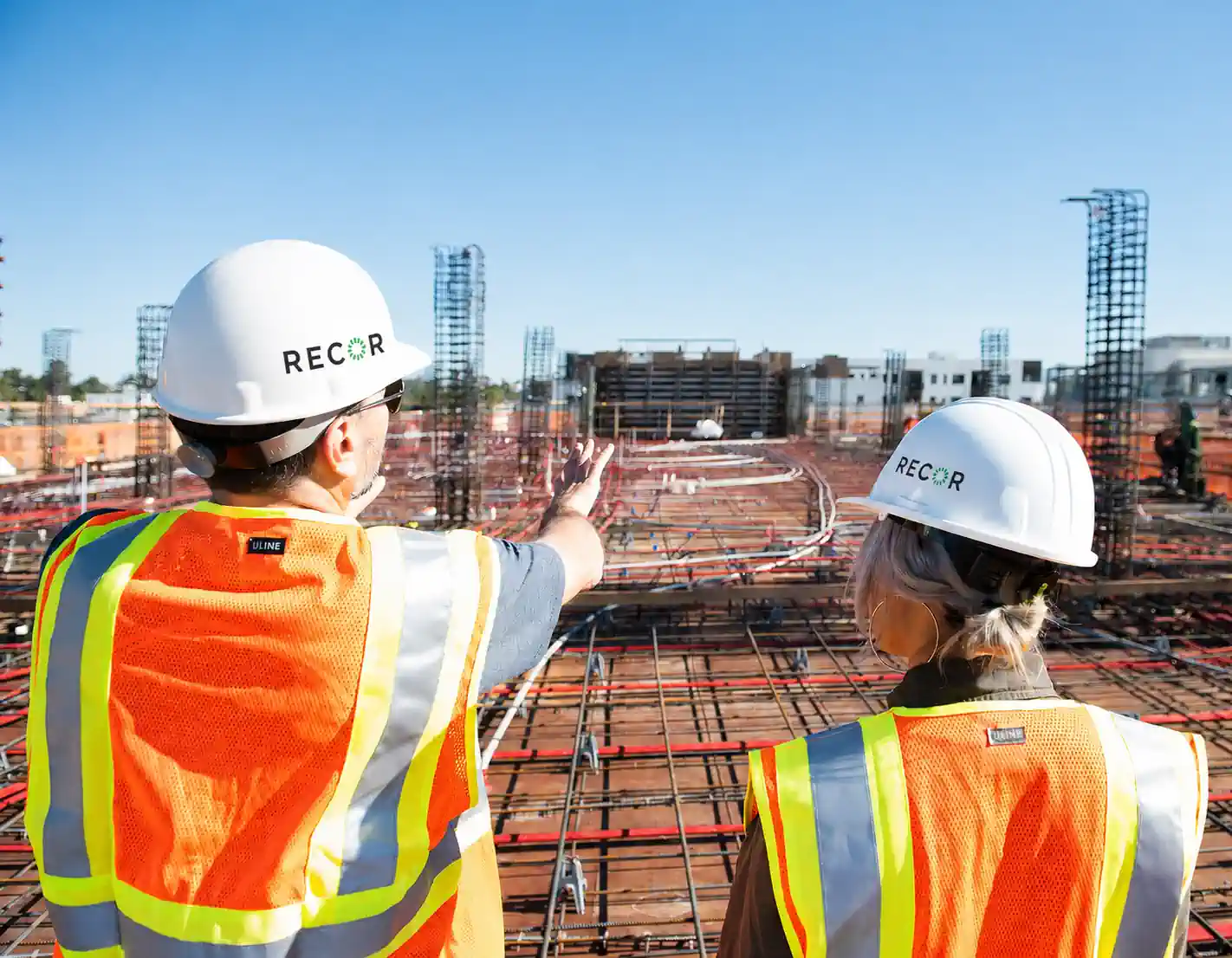 Two RECOR team members in orange hi-vis vests and RECOR-branded white hardhats overlooking a rebar slab with concrete columns rising, one pointing across the site