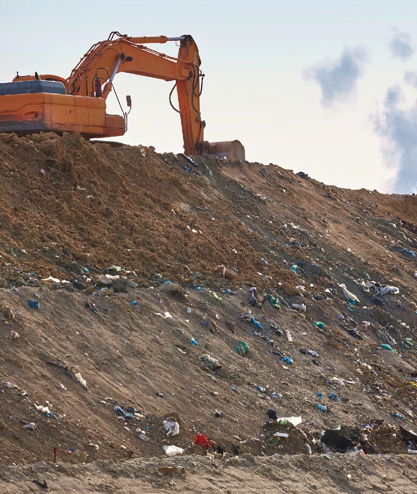 Orange excavator working on top of the Bonnick Road landfill cell capping the slope of dark earth, with scattered waste visible on the embankment and a blue sky behind